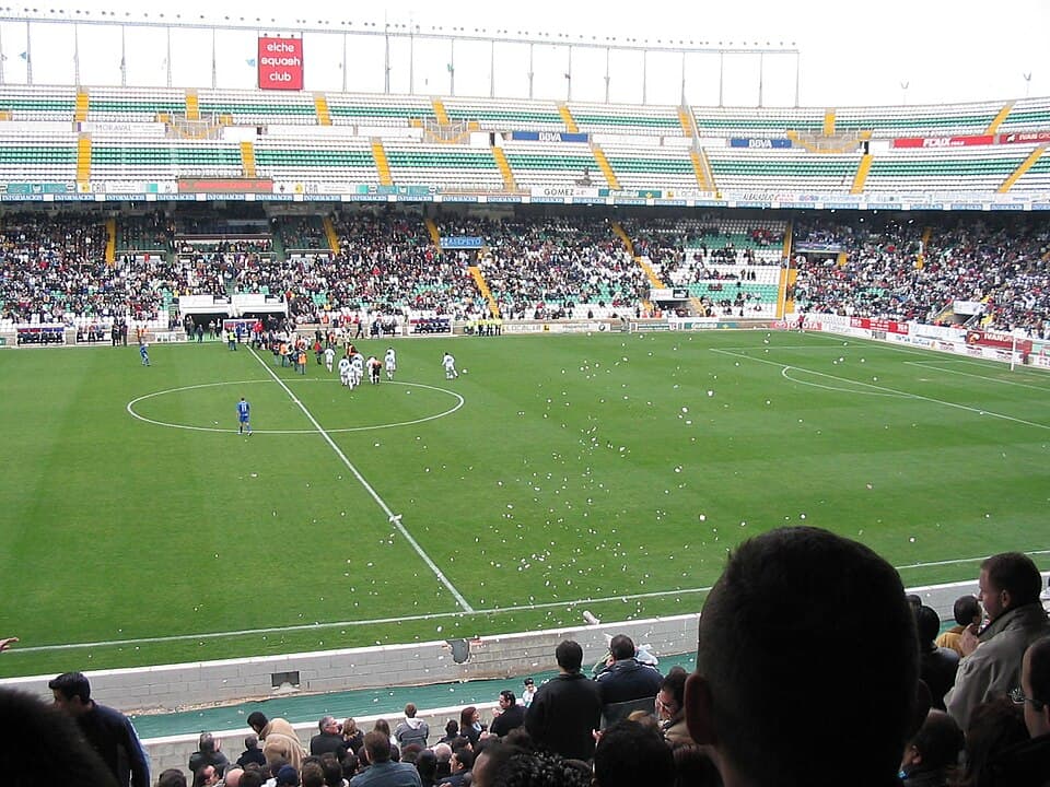 General view of a football stadium packed with fans before kickoff, with the pitch freshly lined and floodlights illuminating the stands.